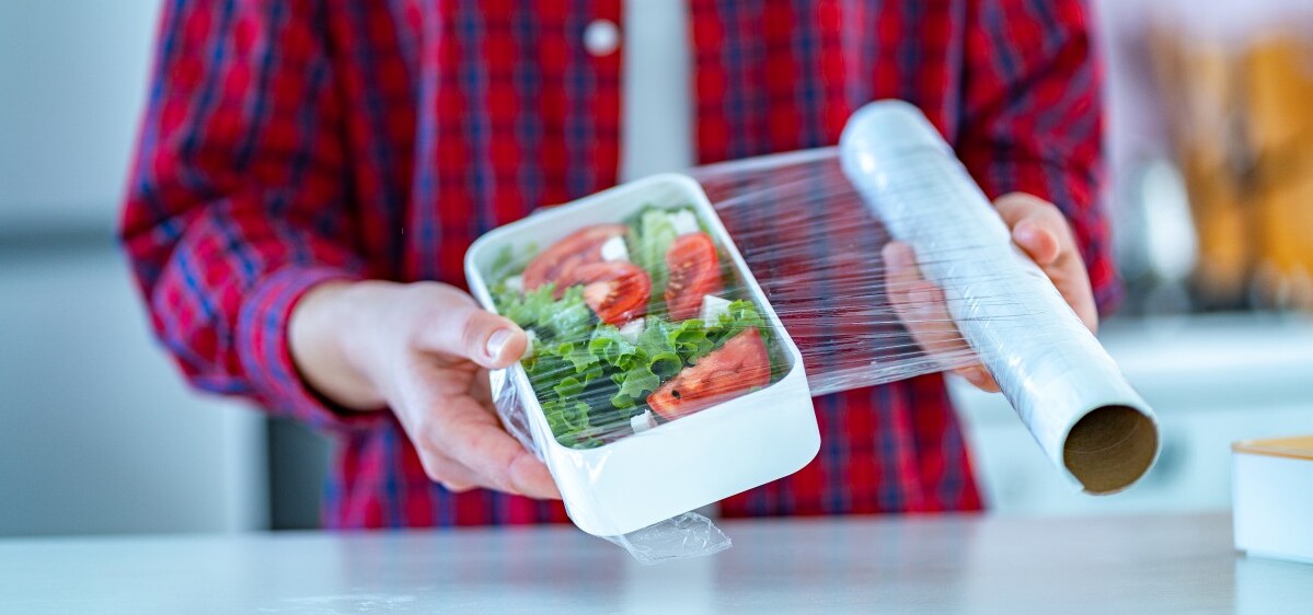a woman is using a cling film in the Kitchen on a plasting trays with salad inside
