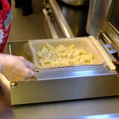 A woman preparing Food-to-gofood packaging in a machine, which seals the top with a flexible film lid